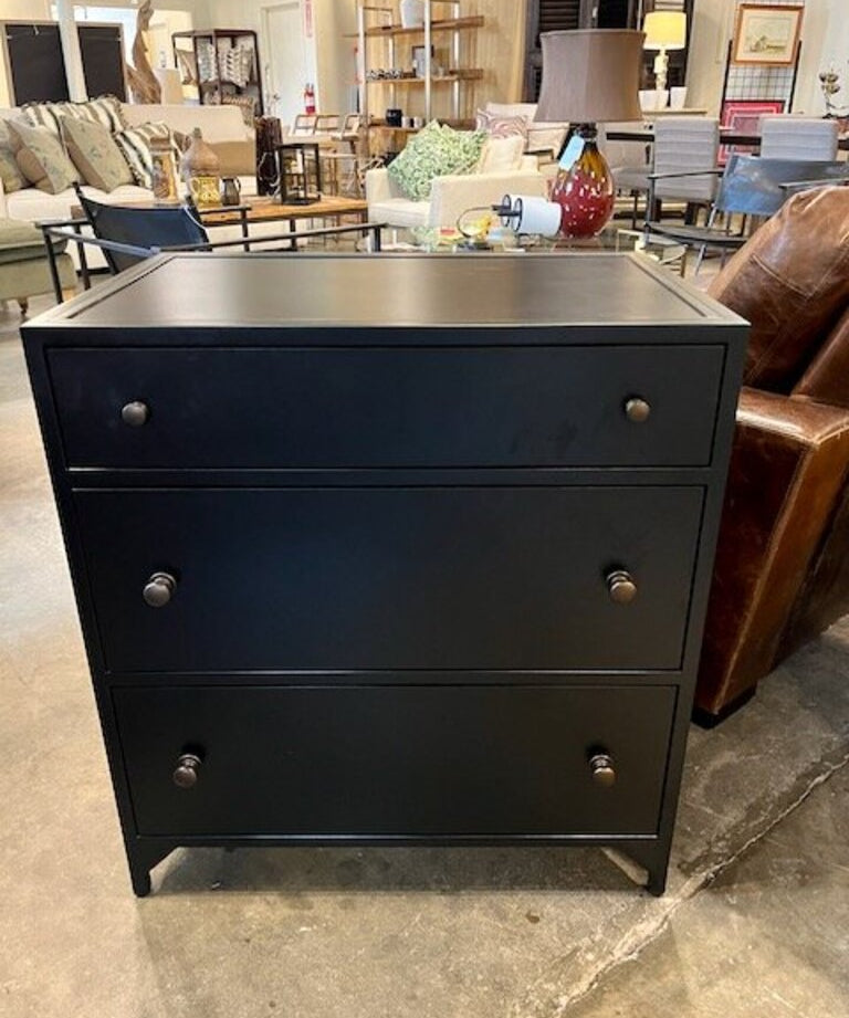Black three-drawer dresser in a furniture store showroom with sofa and lamp background